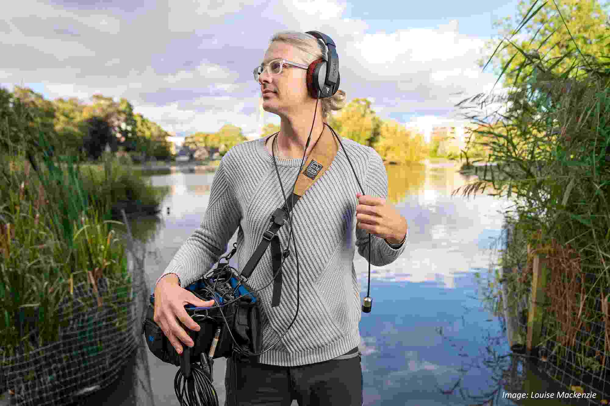 A photograph of the author, David, wearing headphones and carrying sound gear, standing in front of a large pond.  There are reeds in the foreground and trees on the far side of the water.